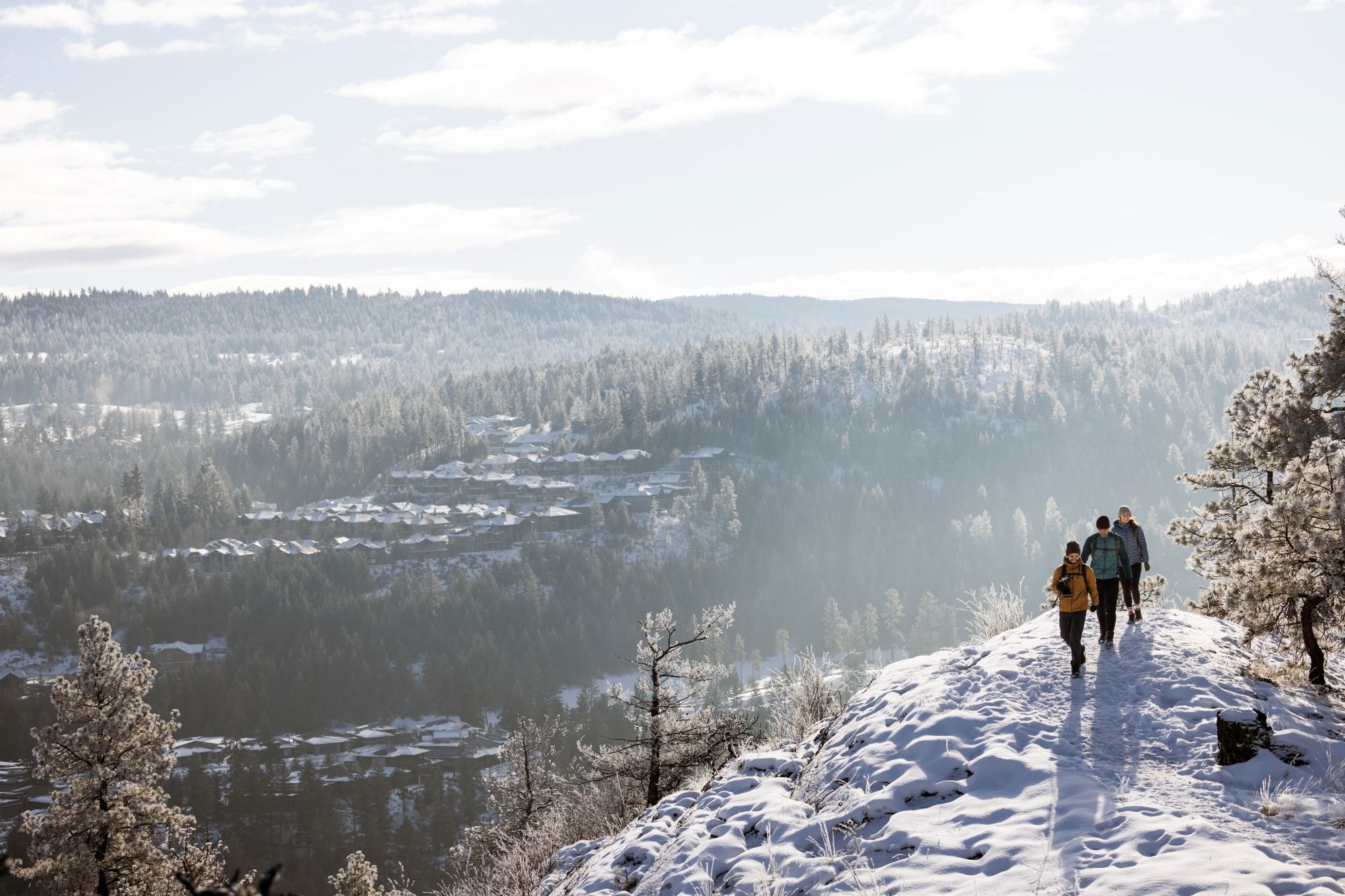 three people walking on snowy ridgeline trail with house views and trees in distance